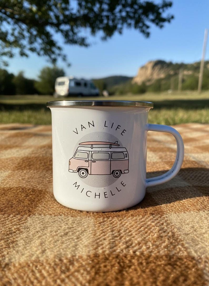 A white enamel mug with a pink campervan illustration and the name Michelle sits on a checkered outdoor blanket.