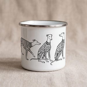 May include: A white enamel mug with a silver-toned rim featuring a black line art illustration of three greyhound-type dogs wearing striped sweaters. The dogs are depicted in various poses, including walking and sitting, against the white surface.