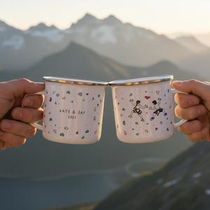 Può includere: Due tazze in smalto bianco con bordi argentati e un motivo di prese da arrampicata colorate. Una tazza riporta la scritta KATE & JAY 2021, l'altra mostra l'illustrazione di due persone che arrampicano con un piccolo cuore rosso sopra.