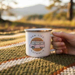 A hand holds a white enamel mug with a pink campervan illustration and the name Nathalie on a patterned blanket outdoors.