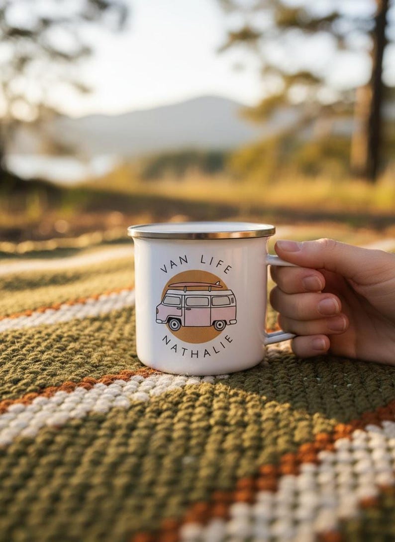 A hand holds a white enamel mug with a pink campervan illustration and the name Nathalie on a patterned blanket outdoors.