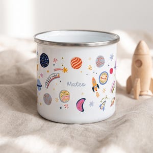 Op de afbeelding: Een witte emaille mok met een zilveren rand voorzien van een kleurrijk ruimtepatroon met planeten, sterren, raketten en de tekst "Space cadet" en "Mateo" op de zijkant. Het ontwerp bevat diverse hemellichamen in oranje, blauw en roze.