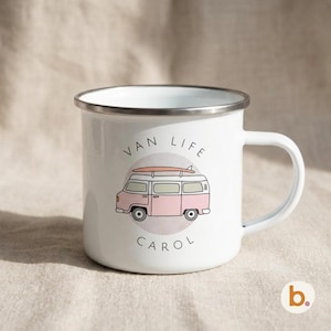 A white enamel mug with a metallic rim is displayed on a beige surface, printed with a pastel pink campervan design and the name CAROL.