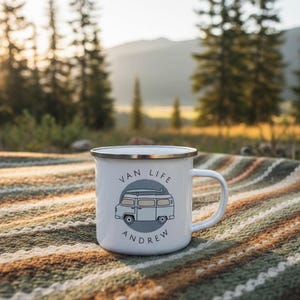 A white enamel mug with a blue campervan illustration and the name Andrew sitting on a textured blanket outdoors.