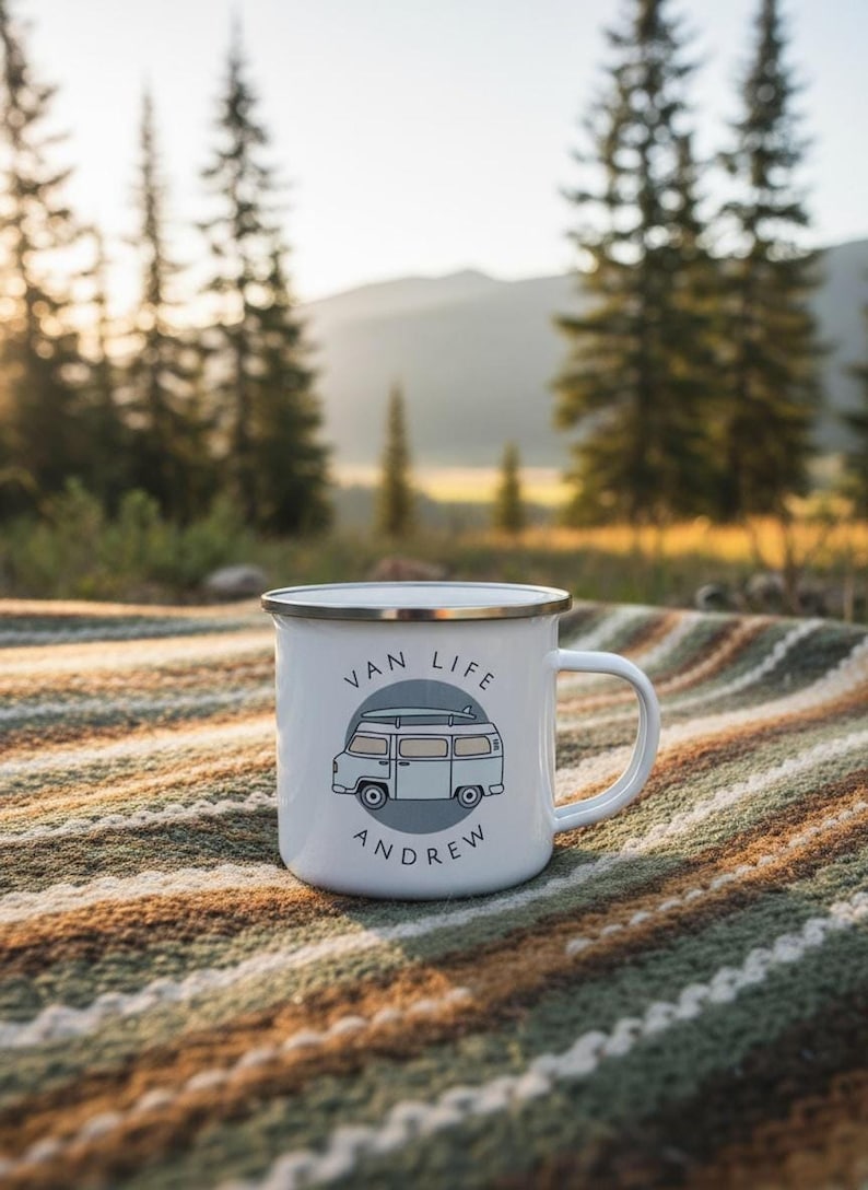 A white enamel mug with a blue campervan illustration and the name Andrew sitting on a textured blanket outdoors.