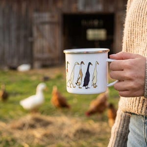 A person wearing a textured beige sweater holds a white enamel duck mug outdoors on a farm, with live ducks and a barn in the background.