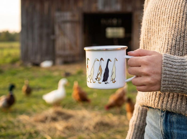 A person wearing a textured beige sweater holds a white enamel duck mug outdoors on a farm, with live ducks and a barn in the background.