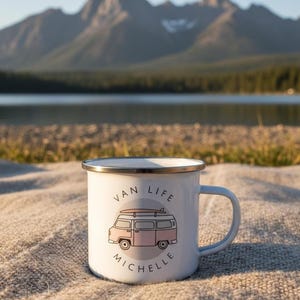 A white enamel mug with a pink campervan illustration and the name Michelle stands on a textured blanket with a lake and mountains in the background.