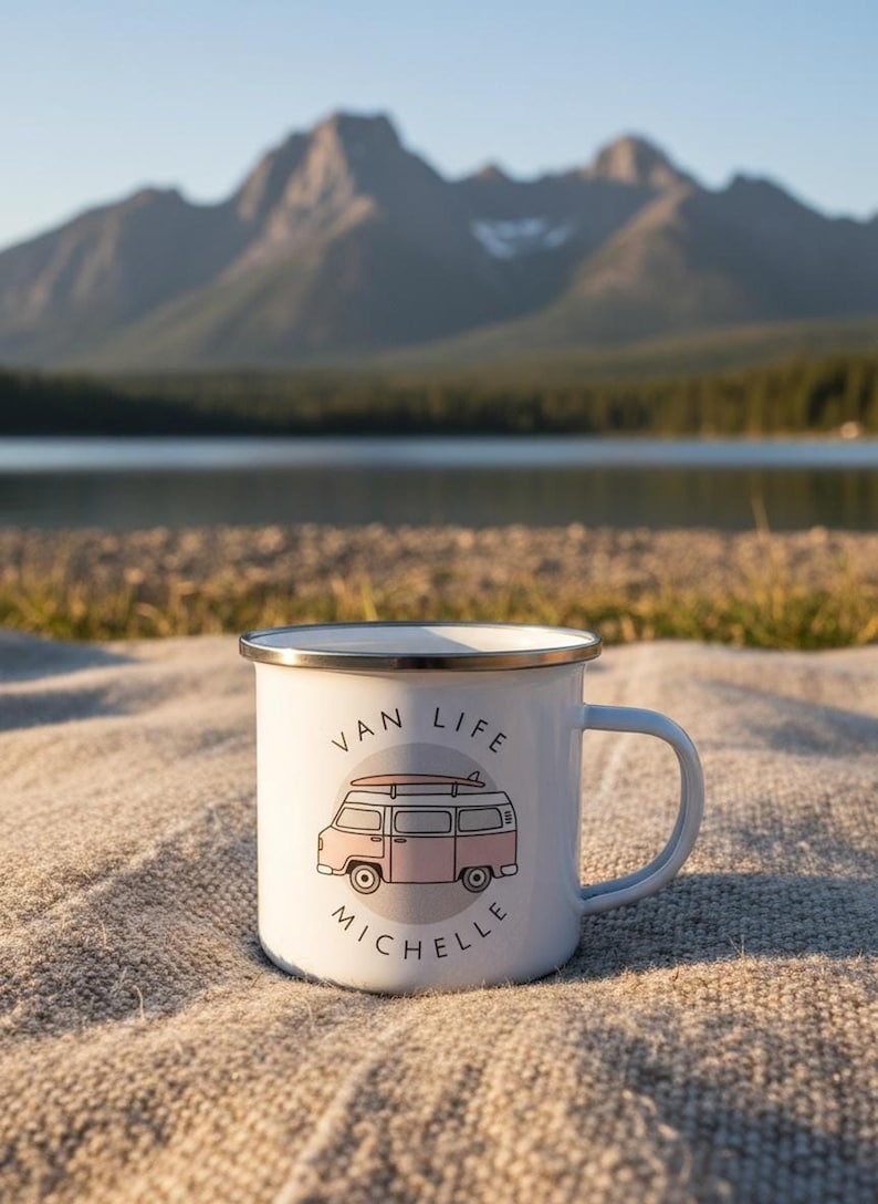 A white enamel mug with a pink campervan illustration and the name Michelle stands on a textured blanket with a lake and mountains in the background.