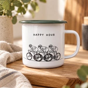 Puede incluir: Una taza de esmalte blanco con un borde y asa de color verde oscuro. La parte frontal presenta un dibujo lineal negro de tres ciclistas pedaleando en fila, bajo el texto negro "HAPPY HOUR" impreso en una fuente sencilla sin remates.