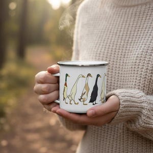 Close-up of a person in a knit sweater holding a steaming white enamel mug printed with runner ducks, with a forest background.