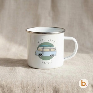 A white enamel mug with a silver rim rests on a textured surface, showing a blue campervan graphic and the text VAN LIFE WOUT.