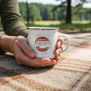 A person holds a white enamel mug featuring a red campervan and the name Claire on a blanket outdoors.