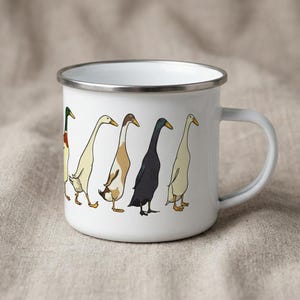 Studio view of a white enamel mug with a silver rim, decorated with a graphic of Indian runner ducks, positioned on a beige cloth.