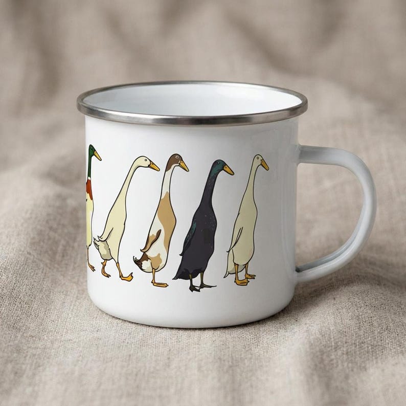 Studio view of a white enamel mug with a silver rim, decorated with a graphic of Indian runner ducks, positioned on a beige cloth.