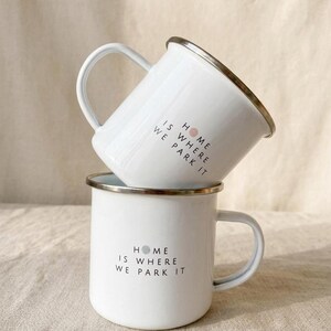 Two white enamel mugs with silver rims are stacked vertically on a neutral cloth surface, both printed with the text HOME IS WHERE WE PARK IT.