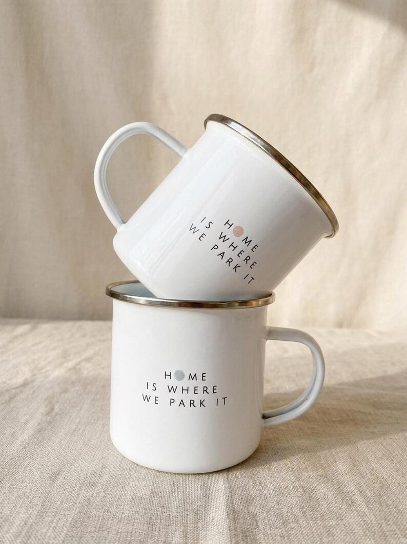 Two white enamel mugs with silver rims are stacked vertically on a neutral cloth surface, both printed with the text HOME IS WHERE WE PARK IT.