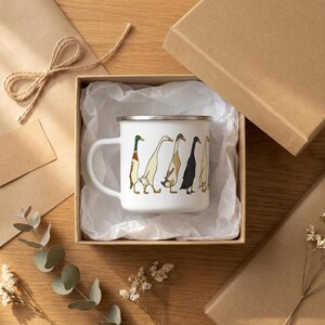 An enamel mug with an illustration of runner ducks sits open inside a cardboard gift box, surrounded by tissue paper, gift wrap, and eucalyptus leaves on a wooden table.