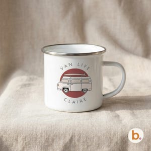 A white enamel mug with a silver rim sits on fabric, displaying a red campervan graphic inside a maroon circle and the name CLAIRE.