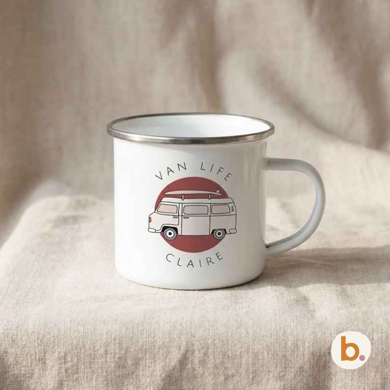 A white enamel mug with a silver rim sits on fabric, displaying a red campervan graphic inside a maroon circle and the name CLAIRE.