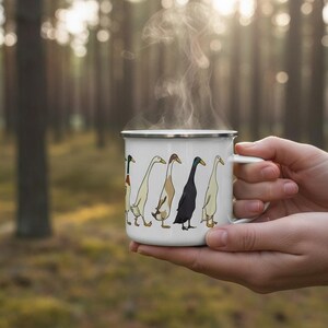 Hands holding a steaming enamel mug with a runner duck graphic against a softly lit pine forest background.