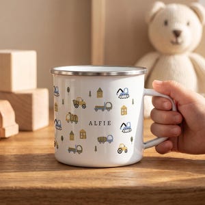 Könnte beinhalten: Ein weißer Emaillebecher mit silbernem Rand und einem wiederkehrenden Muster aus kleinen Baufahrzeugen, Bäumen und Häusern in gedeckten Blau-, Grün- und Gelbtönen. Der Name ALFIE ist mittig in schwarzen Großbuchstaben aufgedruckt.