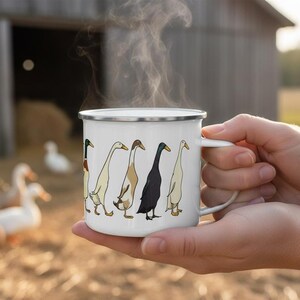 Two hands hold a steaming white enamel mug with a duck illustration, with ducks and a rustic barn visible in a blurred background.