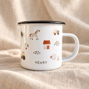 Pode incluir: Uma caneca de esmalte branco com borda e alça pretas, apresentando um padrão de tema de fazenda com um cavalo, uma casa, um caminhão, um trator, ovelhas e árvores. O nome HENRY está impresso em letras maiúsculas pretas na frente.