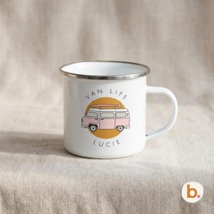 A white enamel mug with a silver rim sits on a neutral fabric background, featuring a pink campervan illustration and the text VAN LIFE LUCIE.
