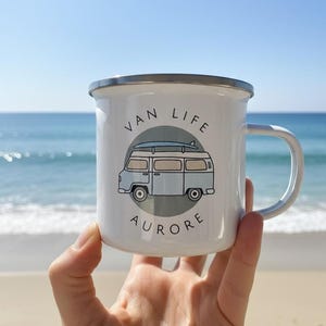 A hand holds up a white enamel mug with a blue campervan illustration and the name Aurore against a beach and ocean backdrop.