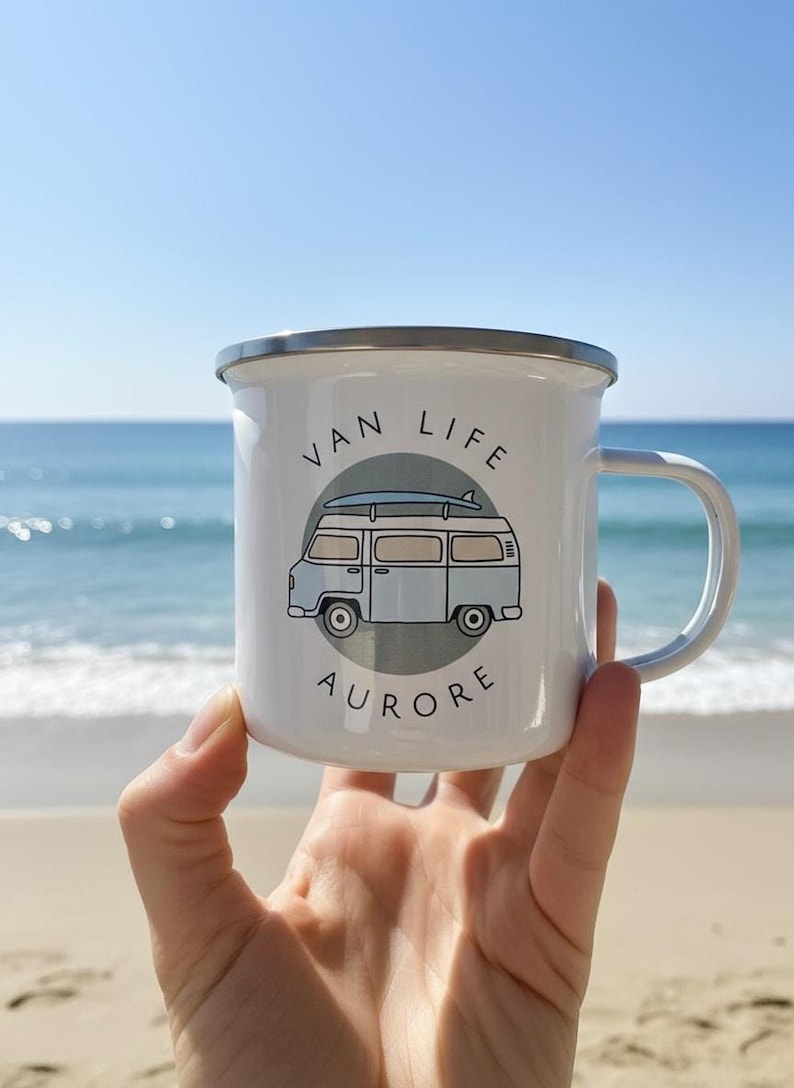 A hand holds up a white enamel mug with a blue campervan illustration and the name Aurore against a beach and ocean backdrop.