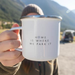 A person holds up a white enamel mug printed with the text Home Is Where We Park It with a mountain landscape in the background.