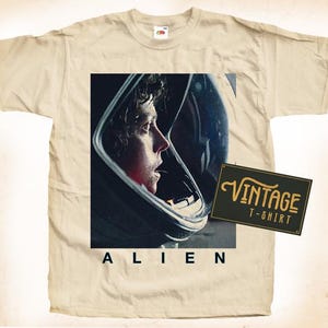 Könnte beinhalten: Beigefarbenes T-Shirt mit einem Grafikmotiv aus dem Film "Alien". Die Grafik zeigt eine Person in einem Weltraumhelm, darunter steht das Wort "ALIEN". Ein Vintage-Etikett lautet "VINTAGE T-SHIRT".