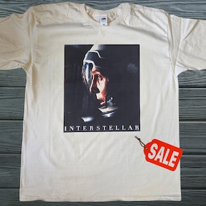 Peut inclure: T-shirt blanc cassé avec un graphisme du film "Interstellar". Le graphisme représente une personne portant un casque spatial. Le mot "INTERSTELLAR" est imprimé en dessous. Une étiquette "SOLDES" rouge est attachée.