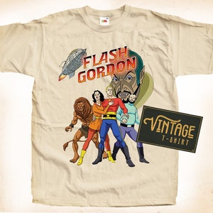 May include: A vintage beige t-shirt featuring a graphic print of Flash Gordon and his crew. The print depicts Flash Gordon, Dale Arden, Dr. Zarkov, and a lion-like creature. The text "FLASH GORDON" is printed in red and yellow lettering. The t-shirt has a vintage label that reads "VINTAGE T-SHIRT".