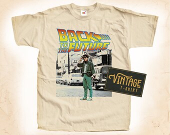 May include: A vintage beige t-shirt with a graphic print of the movie "Back to the Future". The graphic features a character from the movie standing in front of a building with the movie title in a colorful font.