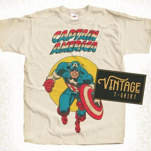 May include: Cream-colored vintage t-shirt featuring a Captain America graphic. The design includes the text "CAPTAIN AMERICA" in red, white, and blue, with an illustration of Captain America holding his shield. A black sign reads "VINTAGE T-SHIRT".