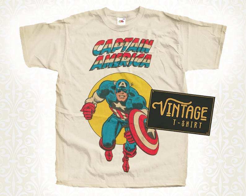 May include: Cream-colored vintage t-shirt featuring a Captain America graphic. The design includes the text "CAPTAIN AMERICA" in red, white, and blue, with an illustration of Captain America holding his shield. A black sign reads "VINTAGE T-SHIRT".