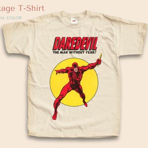 Peut inclure: T-shirt vintage de couleur crème avec un graphisme de Daredevil en costume rouge, posant devant un cercle jaune. Le texte "DAREDEVIL" en rouge et "THE MAN WITHOUT FEAR!" est au-dessus du personnage. Le t-shirt est étiqueté "NATURAL COLOR".