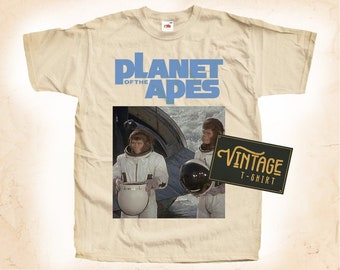 May include: Beige t-shirt featuring the text "PLANET OF THE APES" in blue. The shirt has a graphic of two apes in space suits. A vintage sign is in the lower right corner.