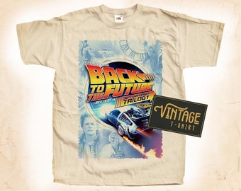 May include: A beige t-shirt with a vintage Back to the Future 30th Anniversary Trilogy graphic print. The print features the DeLorean time machine, Marty McFly, and Doc Brown. The text "Back to the Future 30th Anniversary Trilogy" is printed in orange, red, and blue. The text "Return to the Future" is printed in white. The t-shirt has a vintage look and feel.