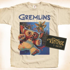 May include: A vintage beige t-shirt with a graphic print of a furry, green, and blue creature from the movie Gremlins. The text "Gremlins" is printed in blue above the image. A black and gold label with the text "Vintage T-Shirt" is in the lower right corner.