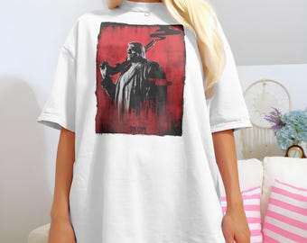 Könnte beinhalten: Weißes Oversize-T-Shirt mit einem Grafikdruck eines Mannes, der eine Waffe hält. Die Grafik ist in Rot, Schwarz und Weiß gehalten und lautet "Sin City".