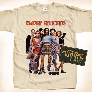 Può includere: Una maglietta vintage beige con una stampa del cast del film Empire Records. La stampa mostra i membri della band in piedi davanti a un boombox con le parole "Empire Records" in un font retrò.