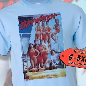 Puede incluir: Una camiseta azul claro con un estampado gráfico del reparto de Baywatch de pie frente a un puesto de salvavidas. El texto "Baywatch" está impreso en rojo y blanco en el gráfico. La camiseta está disponible en tallas S-5XL y está hecha de 100% algodón.