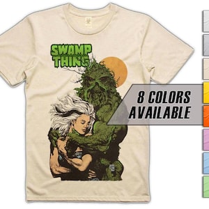 Może przedstawiać: Beżowy t-shirt z krótkim rękawem z grafiką przedstawiającą postać Swamp Thing obejmującą osobę z długimi białymi włosami. Nad postaciami widnieje zielony napis SWAMP THING, a w tle znajduje się żółte słońce.