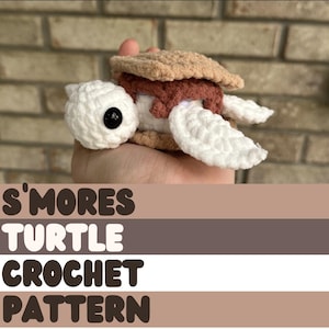 May include: A crocheted turtle amigurumi toy shaped like a s'more. The turtle is white with brown and pink details. The toy is being held in a hand. The text "S'mores Turtle Crochet Pattern" is below the image.