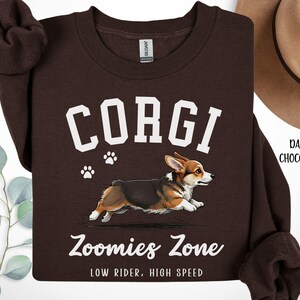 May include: Dark chocolate brown sweatshirt featuring a running corgi illustration with the word "CORGI" in white block letters. Below the corgi is the text "Zoomies Zone" and "LOW RIDER, HIGH SPEED". Two white paw prints are above the corgi.
