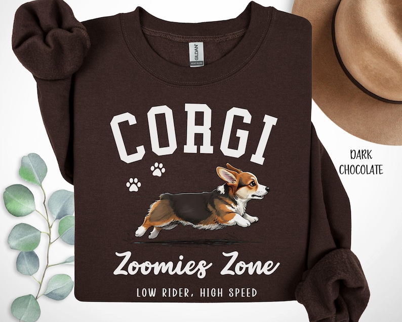 May include: Dark chocolate brown sweatshirt featuring a running corgi illustration with the word "CORGI" in white block letters. Below the corgi is the text "Zoomies Zone" and "LOW RIDER, HIGH SPEED". Two white paw prints are above the corgi.