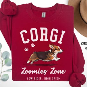 May include: Cardinal red sweatshirt with the word "CORGI" in white block letters, a graphic of a running corgi, and the text "Zoomies Zone: Low Rider, High Speed". The sweatshirt has a crew neck and long sleeves.